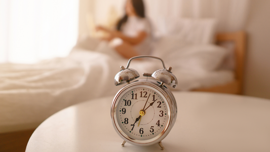 A silver alarm clock is placed on a table while a woman sits up in her bed in the background