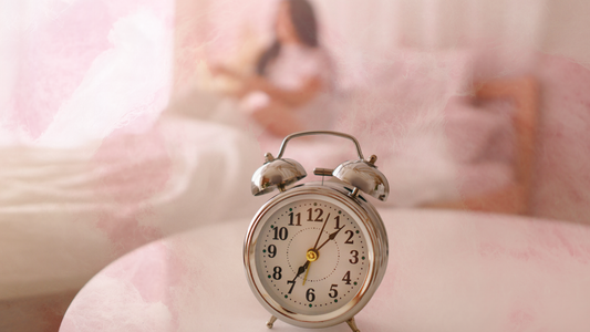 An alarm clock sits on a table in the from of the image, while a person is laying down in the background on a bed.
